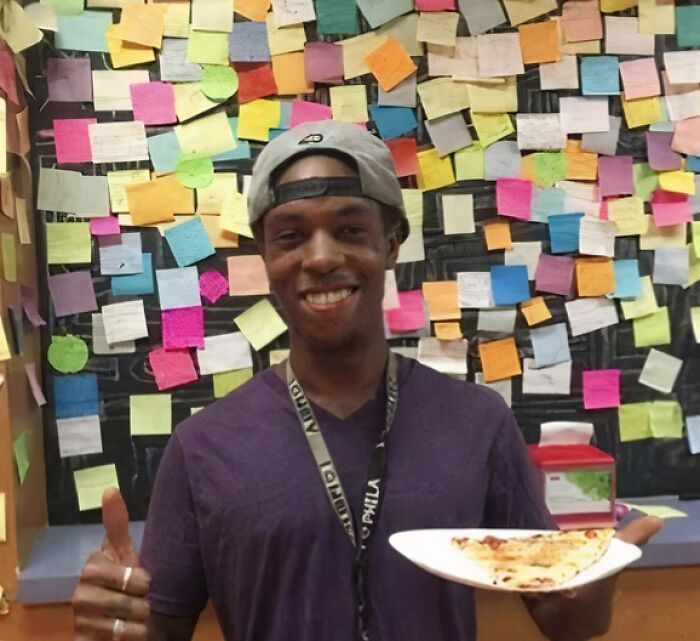 Young man smiling with a slice of pizza and thumbs up in front of a wall covered in colorful heartwarming posts.