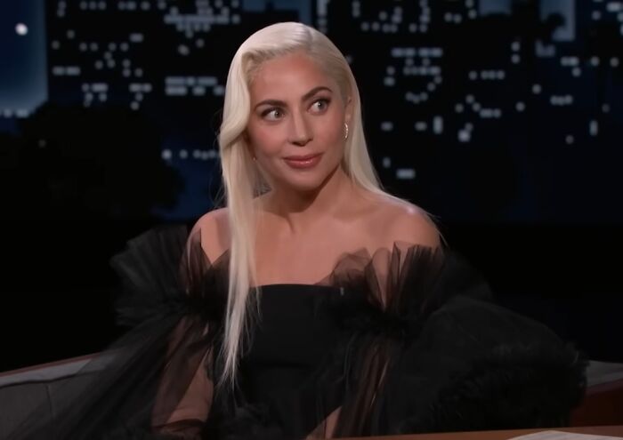 Celebrity with platinum blonde hair in a black outfit appearing on a late-night talk show discussing work and life entitlement.