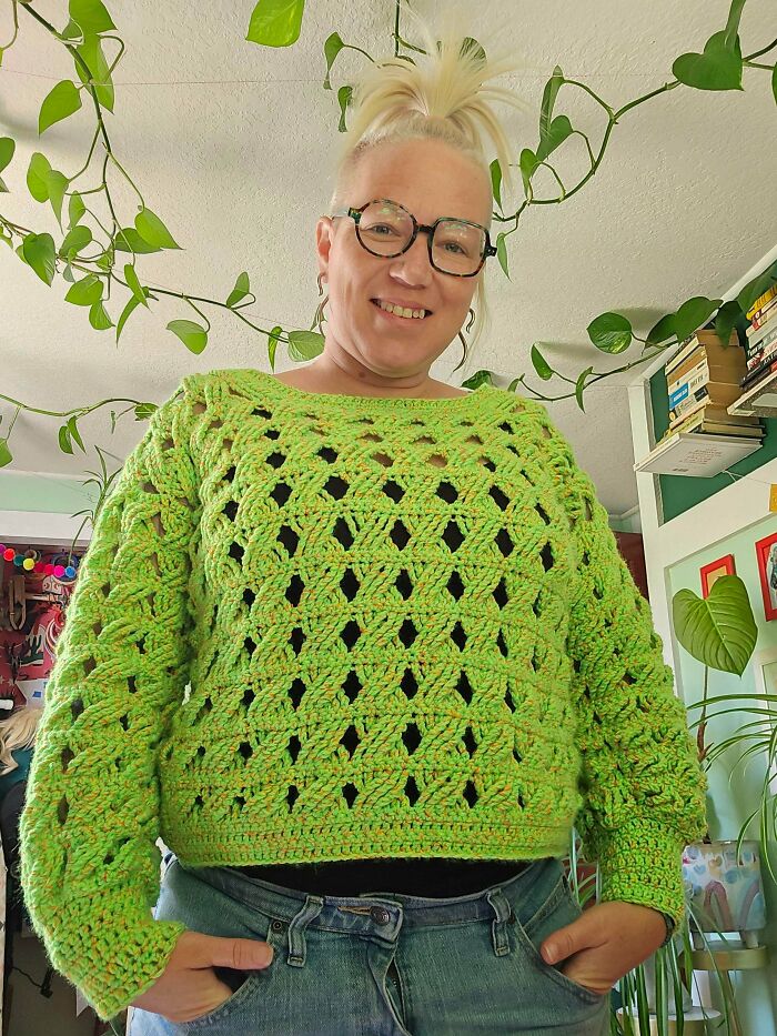Person smiling wearing a handmade bright green crocheted sweater surrounded by indoor plants, showcasing crocheting skills.