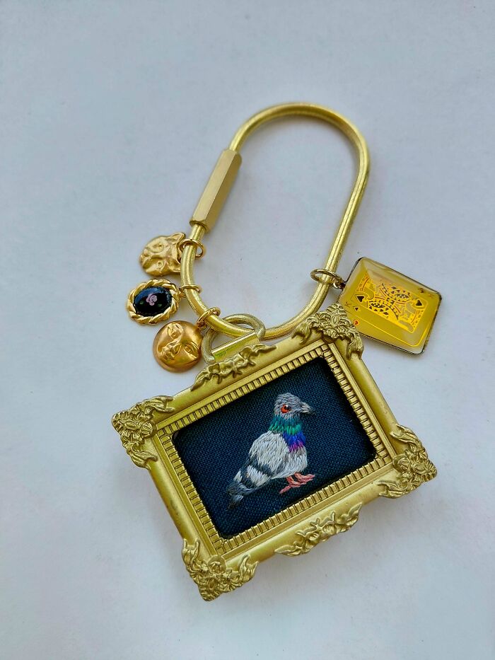 A golden keychain featuring a miniature framed embroidery creation of a detailed pigeon, a stunning embroidery piece.