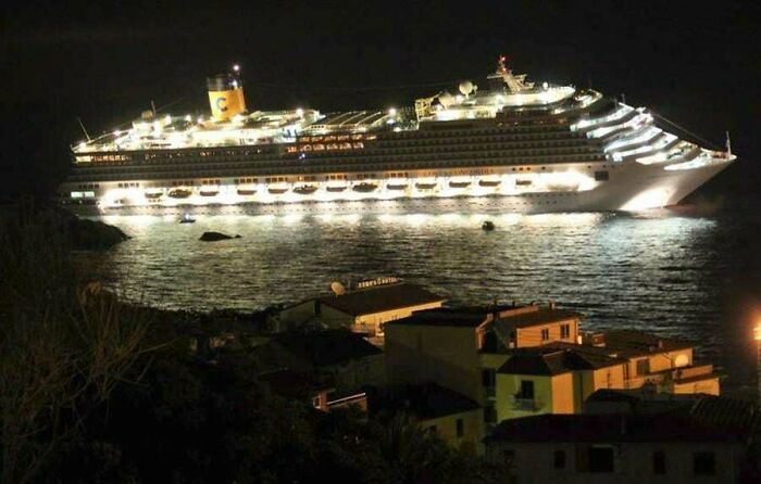 Extremely large things: A giant cruise ship, brightly lit at night, looms over small coastal houses, looking scary and eerie.
