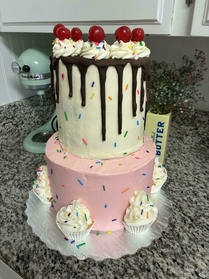 Two-tiered cake with chocolate drip, whipped cream, cherries, and colorful sprinkles, surrounded by mini cupcakes, baked goods display.