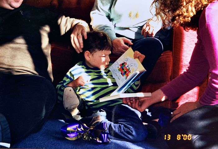 Child in striped shirt reading a colorful book with family members nearby in a warm accidental renaissance moment.