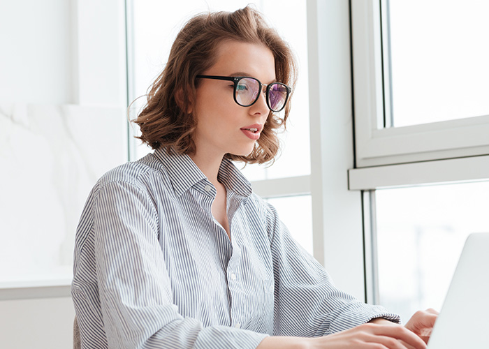 Young woman with glasses working on laptop by window, illustrating moments rich people realize normal life differs widely. - 3