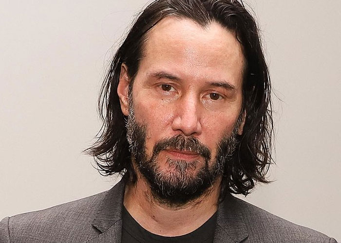 Keanu Reeves, a total gentleman celebrity, posing with a beard and long hair in a gray blazer.