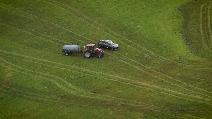 Aerial view of a farmer&rsquo;s tractor spraying manure on a grassy field near a parked black car.