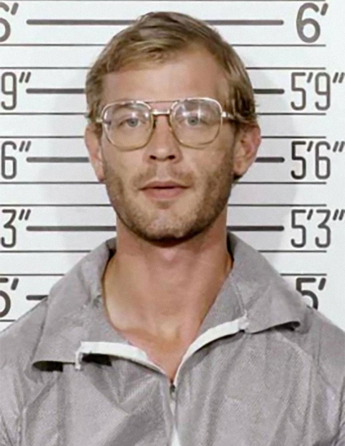 Mugshot photo of a man with glasses and casual jacket in front of a height chart, part of family flexes collection.