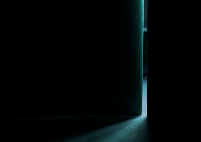 Dimly lit scene showing a slightly open door with light spilling through, evoking security camera footage at night.