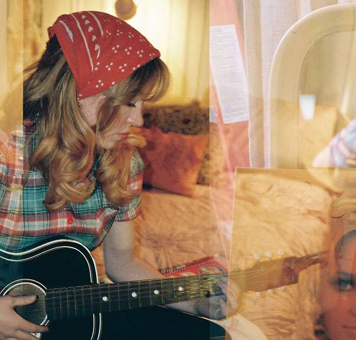 Sydney Sweeney playing guitar indoors with a red bandana and plaid shirt, reflecting a vintage, cozy atmosphere.