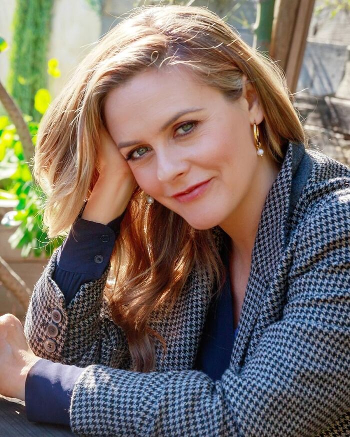 Alicia Silverstone smiling outdoors, wearing a patterned blazer and gold hoop earrings in natural daylight. Alicia Silverstone smiling outdoors, wearing a patterned blazer and gold hoop earrings in natural daylight.