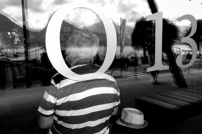Black and white street photograph capturing an unposed moment of a man in a striped shirt through reflective glass.
