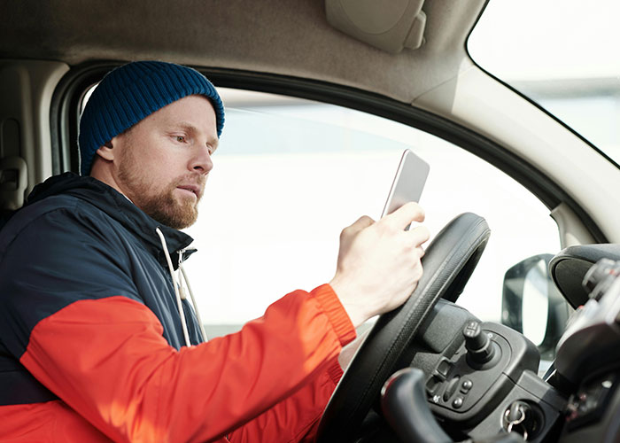 Man wearing a beanie and jacket driving while distracted by looking at his phone, illustrating illegal driving behavior.