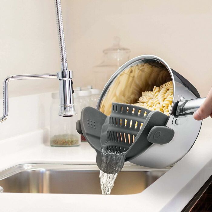 Smart kitchen tool straining pasta water from pot with built-in grey silicone strainer over sink.