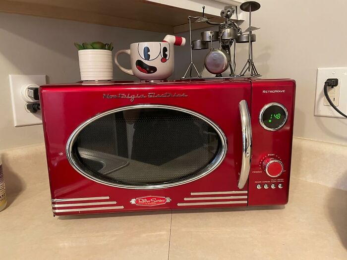 Retro red microwave on a dorm countertop with a cartoon mug, small plant, and metal drum set decoration on top.