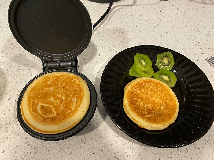 Small electric griddle cooking a pancake next to a black plate with pancake and sliced kiwi in a dorm kitchen setup