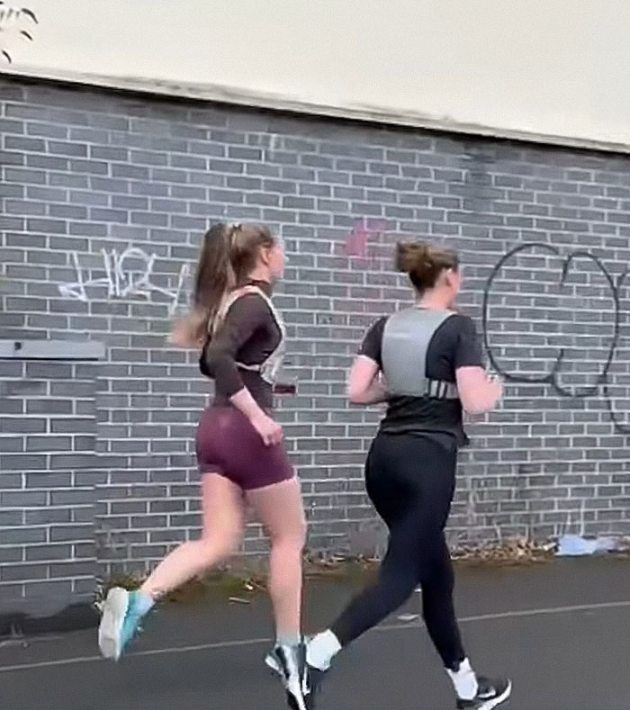 Two women cops wearing running clothes jogging outdoors near a graffiti-covered brick wall in a social experiment.