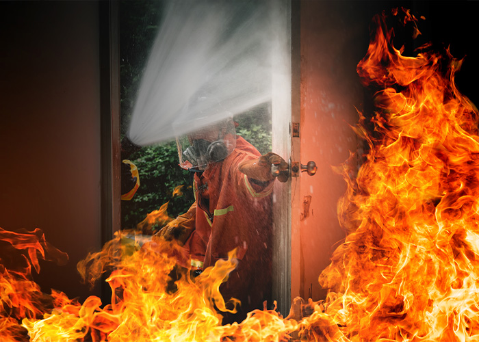 Firefighter in protective gear spraying water to extinguish intense house fire with large flames inside a burning room.