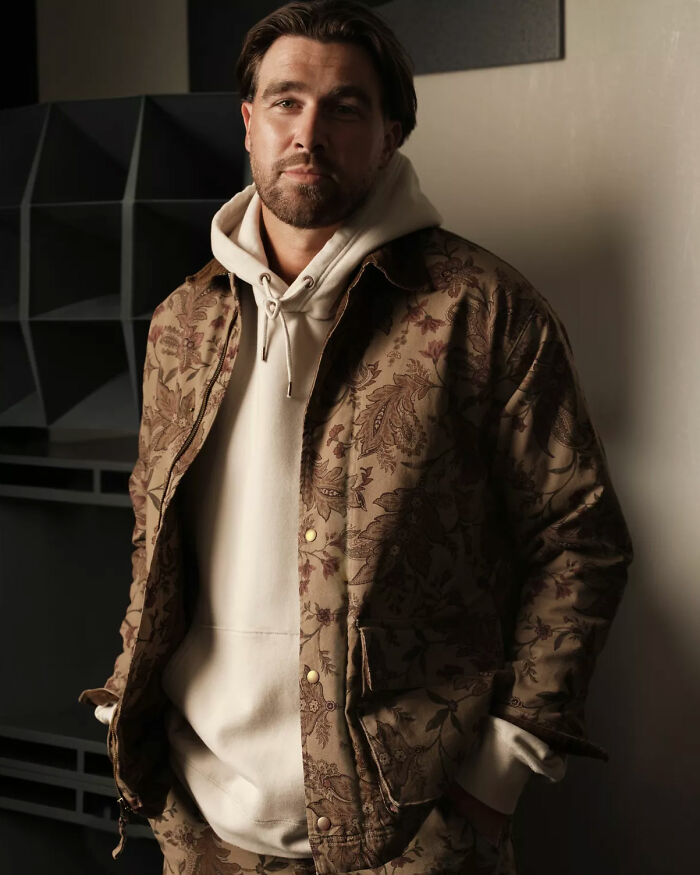 Man wearing beige floral jacket and hoodie posing indoors, linked to fans calling Taylor Swift's engagement a marketing stunt.