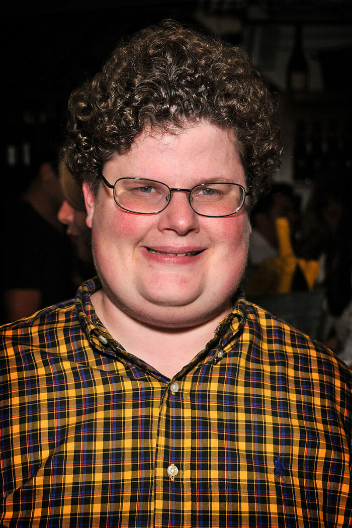Man with curly hair and glasses smiling indoors, wearing a yellow and black plaid shirt, representing new facts and learning keywords.