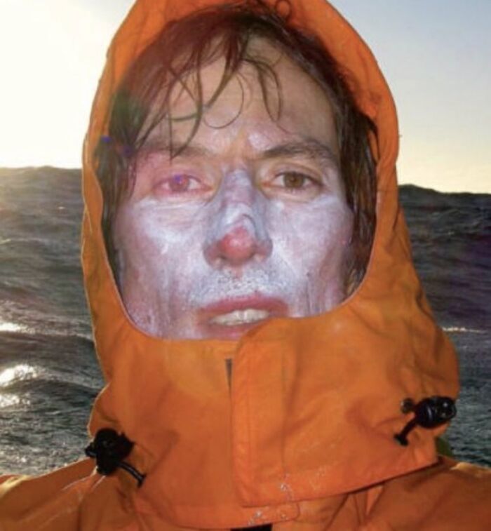 Person with pale, possibly discolored face wearing an orange jacket against a dark ocean background, unsettling image.