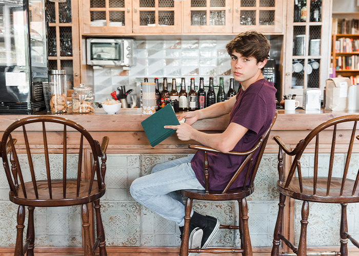 Young man sitting at a bar counter holding a book, surrounded by bottles, illustrating moments rich people realize their normal life. - 7