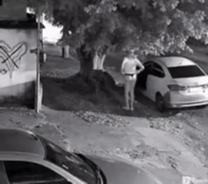 Black and white image showing a person in a thong and wig standing near a car and a tree on a street at night. Black and white image showing a person in a thong and wig standing near a car and a tree on a street at night.
