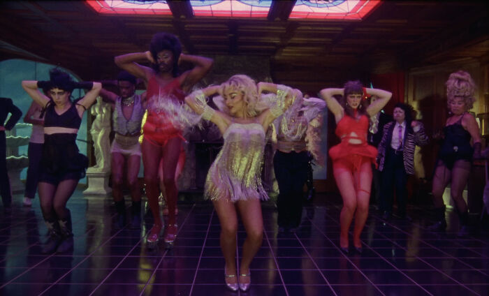 Sabrina Carpenter dancing in a glamorous outfit with backup dancers in a vintage-styled room with dark flooring.