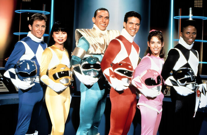 Cast of Power Rangers in colorful costumes holding helmets, evoking nostalgic 90s memories and simpler times.