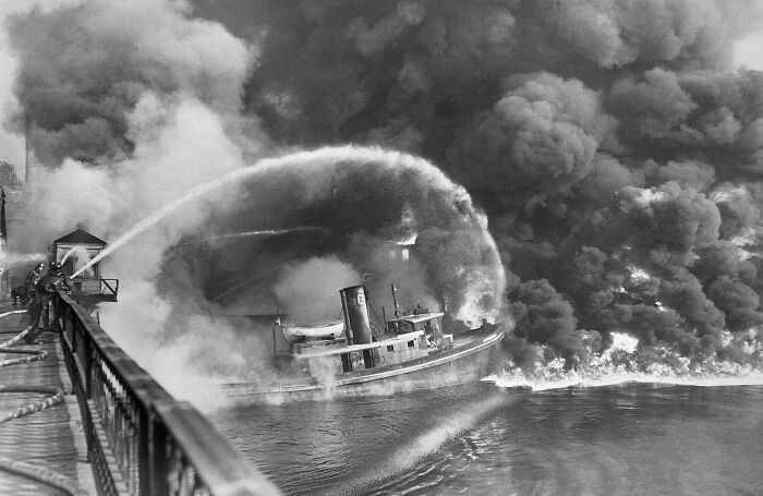 Black and white historical photo of firefighters battling a large fire on a ship with heavy smoke billowing into the sky.