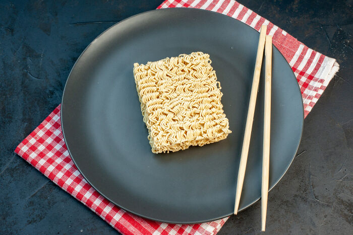 Uncooked instant ramen noodle block on a black plate with chopsticks, showcasing simple confidence in a school kid snack.