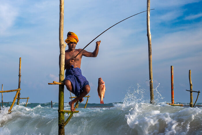 Fisherman in traditional attire catching fish on stilts above ocean waves, showcasing the beauty of cultures.