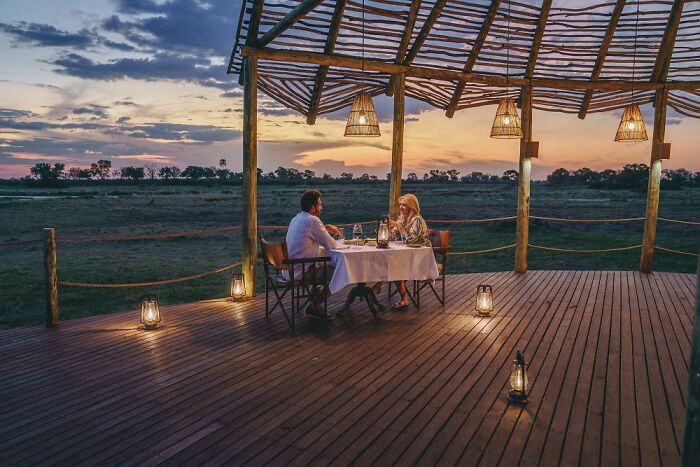 Couple dining at sunset under a rustic wooden shelter, enjoying the serene beauty of the Okavango Delta wilderness.