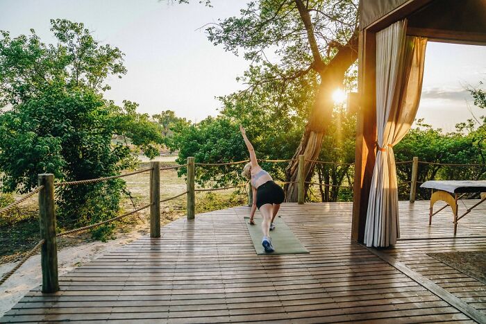Woman practicing yoga on a wooden deck surrounded by lush greenery at the Okavango Delta during sunset.