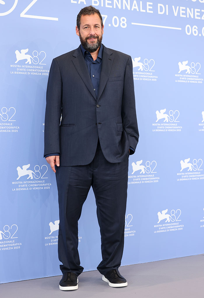 Actor Adam Sandler wearing a dark suit and sneakers, posing at the Venice Film Festival 2025 red carpet event.