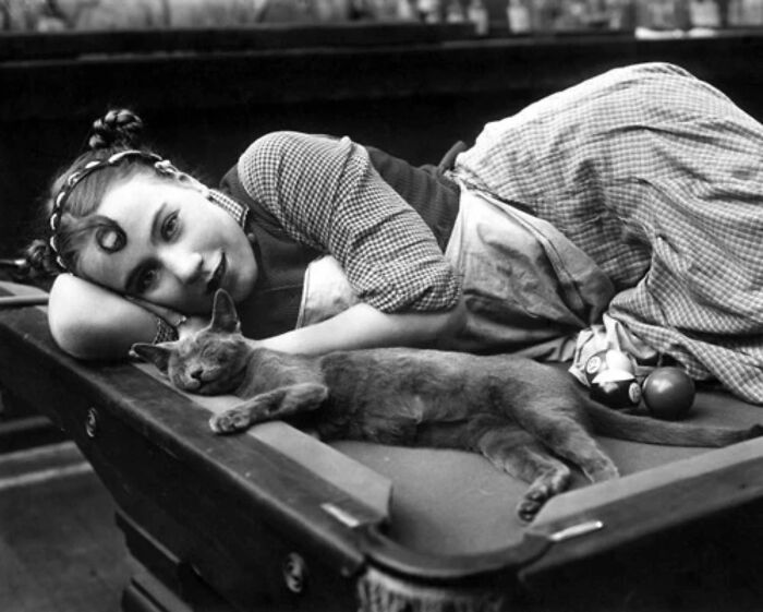 Vintage cat photo showing a woman in period clothing lying beside a sleeping cat on a billiard table.
