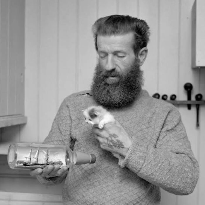 Bearded man in vintage photo holding a small kitten and a ship in a bottle, capturing vintage cat charm.