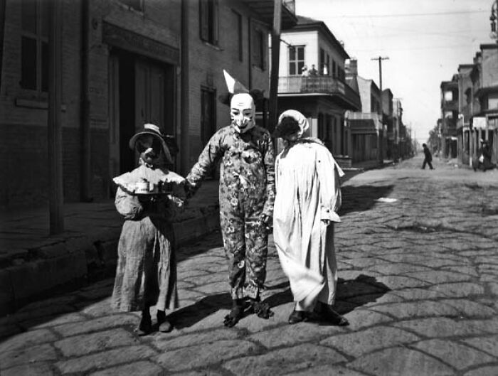 Three people in vintage weird costumes and masks standing on a cobblestone street in a historical photo.