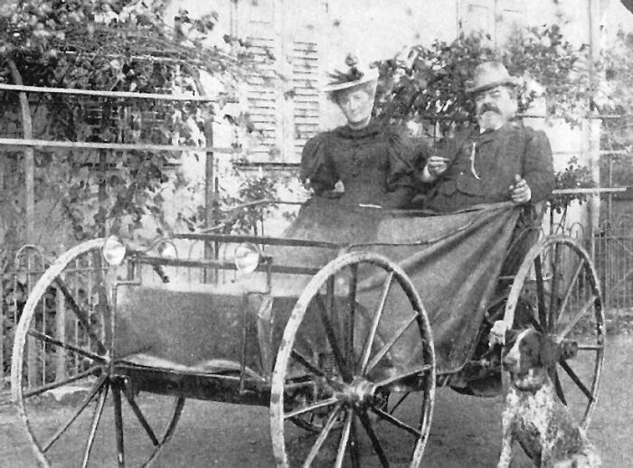 Early 1900s wild cars with large spoked wheels and two passengers dressed in period clothing outdoors.