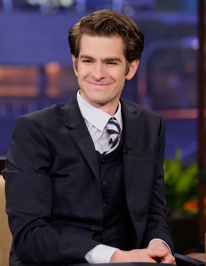 Andrew Garfield in a dark suit and striped tie, smiling during a television interview about his latest look and botox buzz.