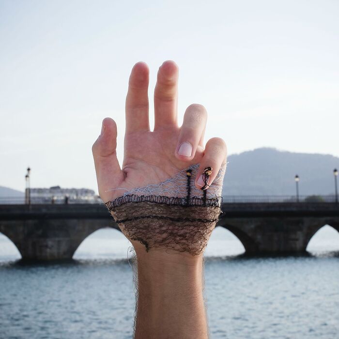 Hand with detailed landscape stitched onto skin, showing mountains and water elements in artistic embroidery.
