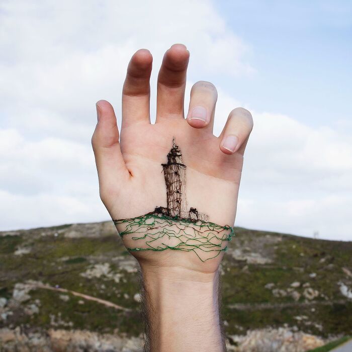 Hand with stitched landscape artwork on skin showing a lighthouse and green hills against a cloudy sky background.