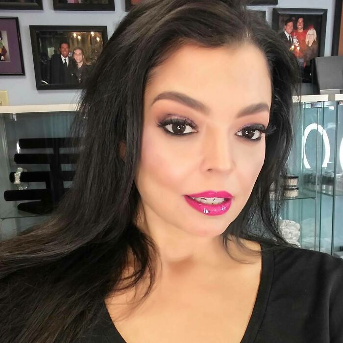Miss Universe winner with long dark hair and pink lipstick posing indoors with framed photos and display case behind her.