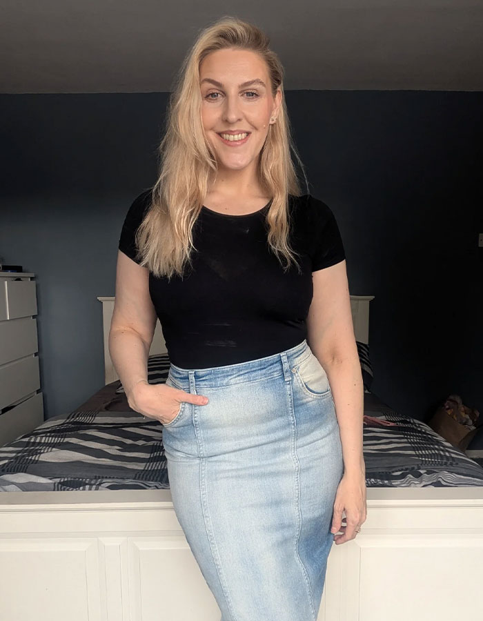 Smiling woman standing in a bedroom wearing a black top and denim skirt, relating to passenger sharing worst experience with child on flight.