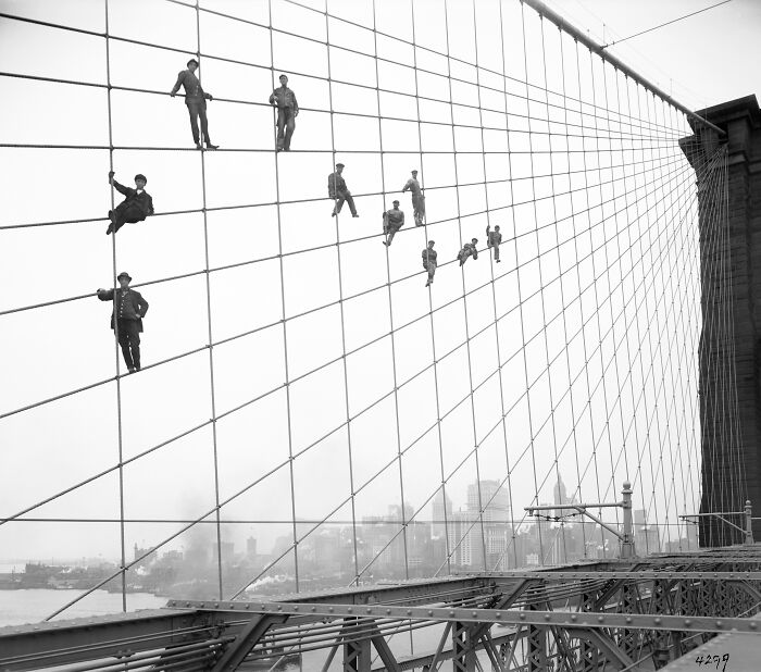Workers sitting on suspension cables of a bridge high above the city in a rare historical photo.