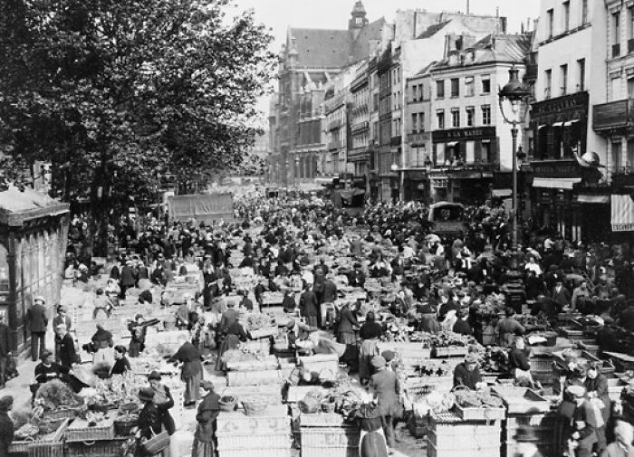 Crowded historical market scene with numerous people and stalls lining a wide city street in a vintage black and white photo.