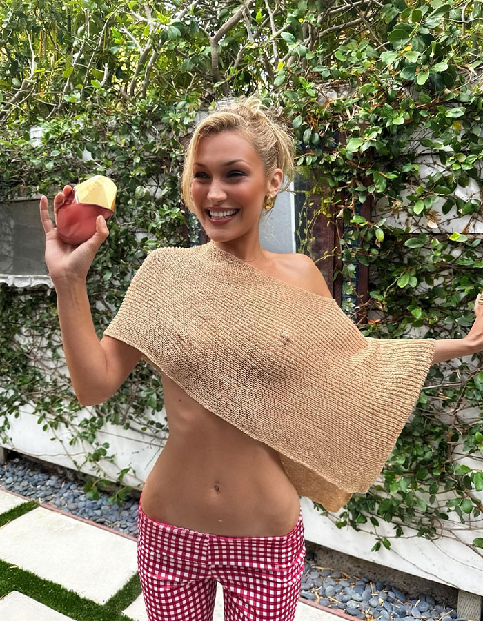 Bella Hadid in a daring braless look holding a perfume bottle outdoors with greenery in the background.