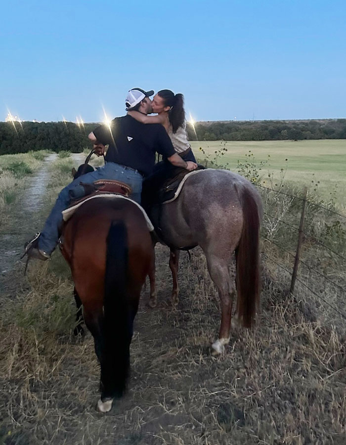 Couple riding horses on a trail at dusk, sharing a kiss, with open fields and clear skies in the background.