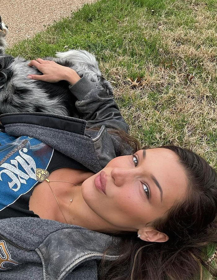 Bella Hadid posing outdoors lying on grass with dog, showing a casual, natural look for new perfume launch.