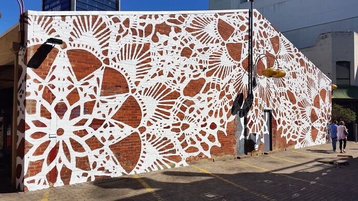 Large street art mural with white intricate patterns on a brick wall under a clear blue sky in an urban setting.