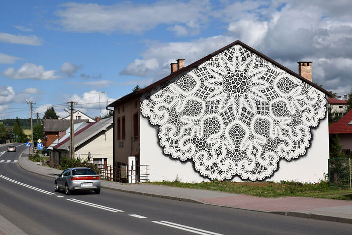 Large intricate lace pattern street art painted on a house wall along a roadside under a partly cloudy sky.
